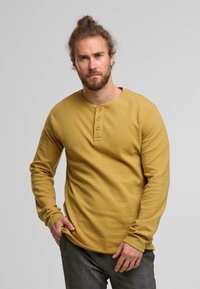 Hombre con barba y moño de pelo, vistiendo una camiseta de manga larga color mostaza y pantalones grises, de pie contra un fondo claro liso.