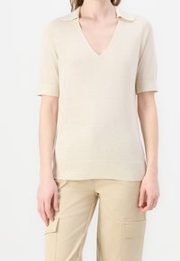 Top in maglia beige a maniche corte con scollatura a V e colletto. Texture liscia con dettagli a coste sulle maniche e sull'orlo. Indossato con pantaloni cargo beige.
