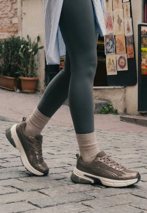 Osoba v šedých legínách, béžových ponožkách a hnědých teniskách kráčí po dlážděné ulici poblíž zdi s uměleckými tisky a květináči.