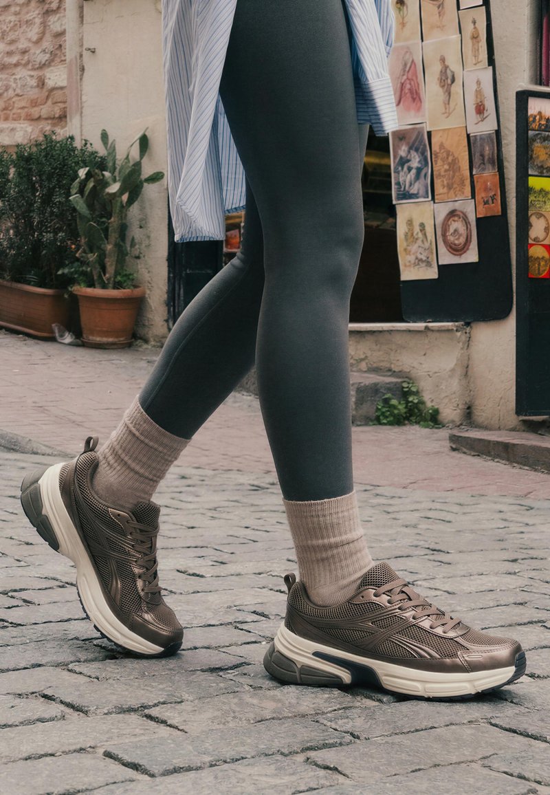 Persona che indossa leggings grigi, calzini beige e scarpe da ginnastica marroni che cammina su una strada lastricata vicino a un muro con stampe artistiche e piante in vaso.