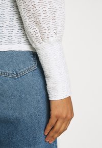 Handen hvilar vid sidan iförd en vit spets långärmad topp och blå denimjeans med synlig sömnad på bakfickorna.