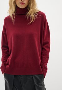 Pull à col roulé bordeaux avec une texture douce et une coupe décontractée. Manches longues et poignets côtelés; porté sur un pantalon en cuir noir.