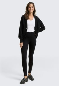 Zwarte geribbelde cardigan met knopen, witte aansluitende t-shirt, en zwarte high-waisted leggings. Zwarte loafers maken de outfit compleet.