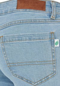 Lichtblauwe denim jeans hebben een bruine leren label aan de tailleband met "URBAN CLASSICS," contrasterende oranje stiksels en een klein groen bladeren label.