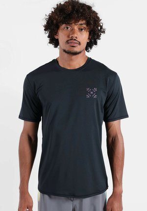 Homme aux cheveux bouclés portant un t-shirt noir à manches courtes avec un petit logo géométrique violet sur le côté gauche de la poitrine.