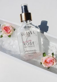 Bouteille en verre transparent avec un capuchon doré, étiquetée "Eau de Rose Bio". Contient un liquide rose, entouré de glace et de fausses roses roses pâles.