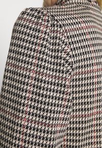 Veste en mélange de laine avec un motif pied-de-poule noir et beige, rehaussée de lignes rouges, présentant une conception d'épaules structurées.