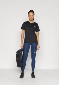 Μαύρο μπλουζάκι και σκούρα μπλε κολάν, και τα δύο με λογότυπα The North Face· το μοντέλο κρατάει μαύρο σακίδιο πλάτης· ύφασμα λείο και εφαρμοστό σχέδιο.