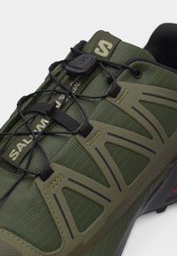 Primo piano di una scarpa da trail running verde con sistema di allacciatura rapida nero e marchio "Salomon Trail" sulla linguetta.