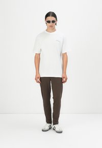 White short-sleeve T-shirt with light blue text, brown slim-fit pants, and black sunglasses. Model wearing white sneakers with black accents.