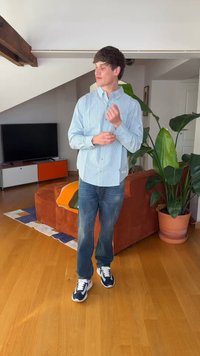 Tânăr bărbat își ajustează manșetele cămășii cu dungi albastre deschise în sufragerie, cu canapea portocalie, plantă mare verde și televizor în fundal.