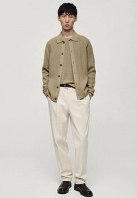Cardigan a maglia beige sopra un maglione beige chiaro, abbinato a pantaloni crema e stivali slip-on marrone scuro. Dettagli con bottoni sul cardigan.