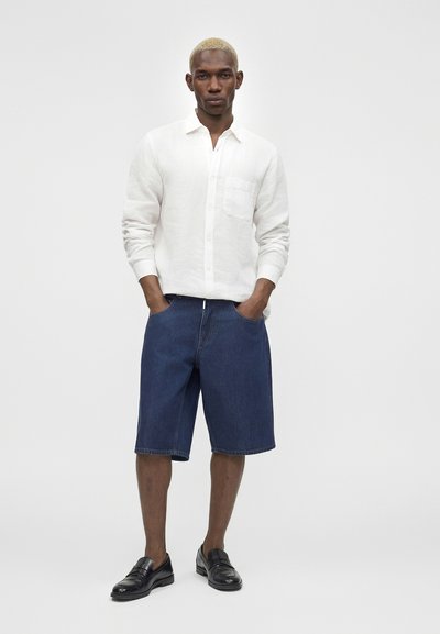 Άνδρας με κοντά ξανθά μαλλιά που φοράει λευκό πουκάμισο με κουμπιά, μπλε τζιν σορτς μέχρι το γόνατο και μαύρα επίσημα παπούτσια, στέκεται με τα χέρια στις τσέπες.