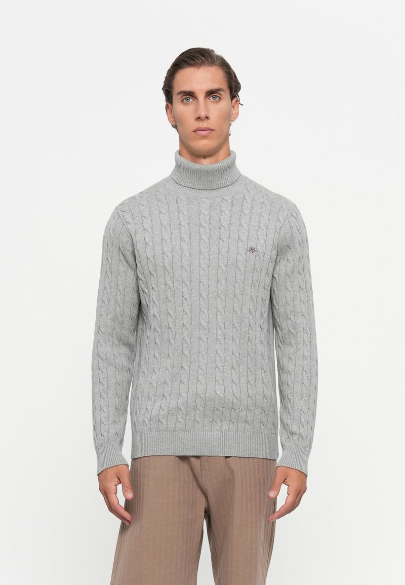 Mann in einem hellgrauen Kabelstrick-Pullover mit Rollkragen und beigen Hosen, der vor einem schlichten weißen Hintergrund steht.
