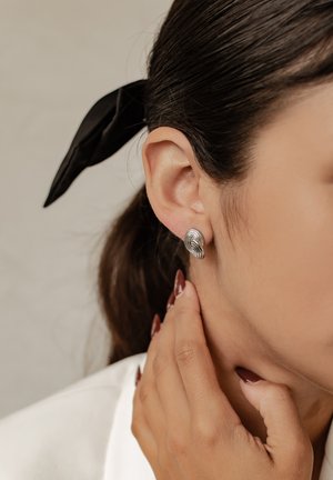 Silberne Ohrringe mit einem wirbelnden Design und glänzender Oberfläche. Das Modell hat dunkles Haar, das mit einem schwarzen Band zusammengebunden ist, und berührt ihren Hals.