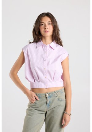 Jonge vrouw met bruin haar draagt een mouwloze lichtroze gestreepte crop top met knopen en lichtblauwe spijkerbroek, staat met één hand in de zak.