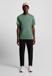 Maglietta verde a maniche corte con un piccolo logo giallo, abbinata a pantaloni neri e sneakers dai toni chiari. Design semplice e dalla vestibilità rilassata.