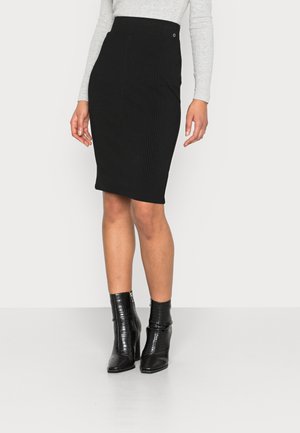 Vrouw draagt een zwarte geribbelde kokerrok, een grijs shirt met lange mouwen en zwarte textuur enkellaarsjes tegen een effen witte achtergrond.