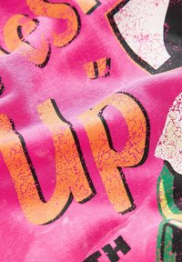 Lyserødt stof med stort slidt tekst i orange og sort. Designet indeholder et grafisk element, der fremhæver det dristige udseende.