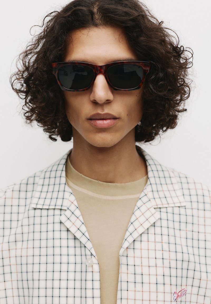 Ochelari de soare cu tipar tartan, cu lentile întunecate, purtați cu o cămașă bej și o cămașă albă cu model în carouri, având accente în bleu deschis și roz.
