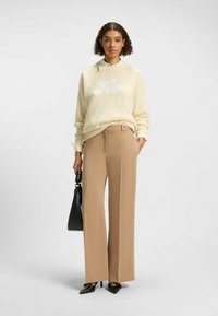 Maglione lavorato a maglia crema con "Hugo" in bianco, abbinato a pantaloni larghi color beige. Una borsa nera e decolleté neri appuntiti completano il look.
