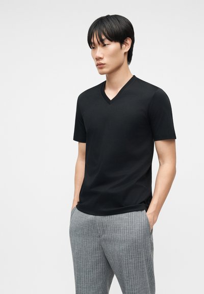 Νέος άνδρας που φοράει μαύρο κοντομάνικο μπλουζάκι με V λαιμόκοψη και γκρι παντελόνι με ρίγες, με τα χέρια στις τσέπες, στέκεται μπροστά σε απλό φόντο.