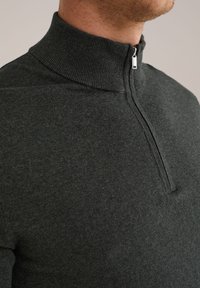 Férfi sötét szürke kötött pulóverben, magas bordás gallérral és elöl fél cipzárral, közeli felvétel a nyakán és vállain.