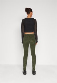 Black cropped knit top with long sleeves, paired with dark green corduroy pants. Model wears black shoes. Plain white background.