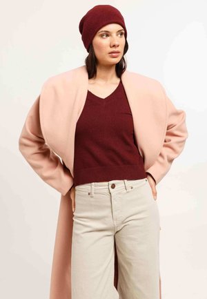 Woman wearing a maroon beanie and sweater, light beige pants, and a pink overcoat draped over her shoulders, standing with hands on hips.