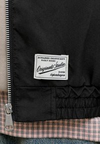 Veste noire avec une étiquette blanche indiquant « Jack & Jones Creative Dept. Originals Studio ©1990 Copenhagen » et une fermeture éclair argentée à côté d'une chemise à carreaux.