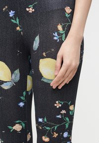 Κοντινό πλάνο ενός χεριού που ακουμπάει πάνω σε μαύρο κολάν με λουλουδάτο και μεγάλο κίτρινο μοτίβο λεμονιού.