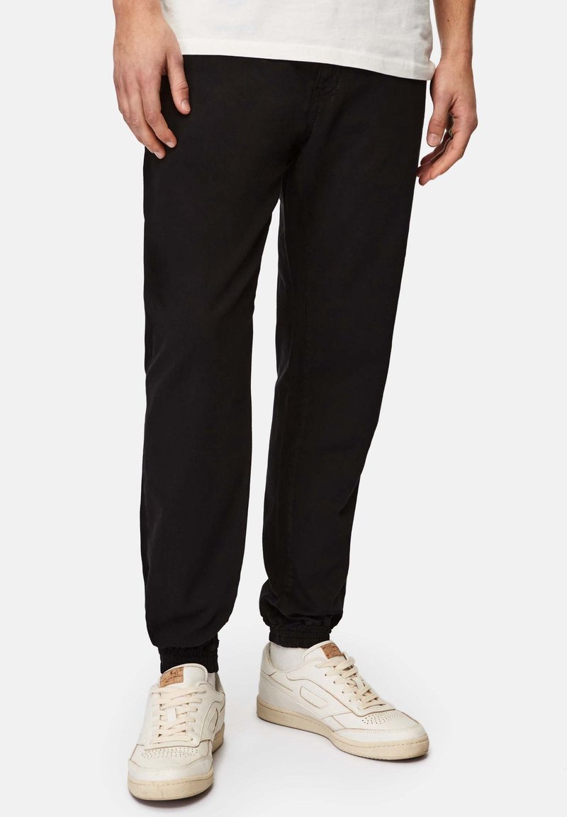 Schwarze Jogginghosen mit elastischen Fußöffnungen, aus Baumwolle gefertigt. Kombiniert mit weißen Sneakern, die über strukturierte Details und ein klassisches Schnürdesign verfügen.