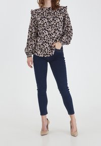 Leopardenmuster-Bluse mit Rüsche an den Schultern, langen Ärmeln und lockerer Passform; kombiniert mit dunkelblauen Skinny-Jeans und beigen High Heels.