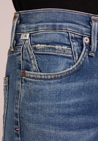 Blue denim jeans with a fitted waist, featuring a distressed finish, five-pocket design, and tan leather patch at the back.