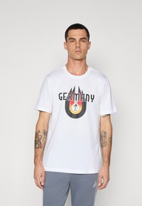 Hvid bomulds t-shirt med et grafisk print, der viser ordet "TYSKLAND" og et cirkulært logo, sat mod en ensfarvet baggrund.