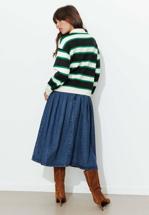 Femme portant un pull rayé vert, noir et blanc, une jupe midi en jean plissée bleue, et des bottines à talons en daim brun, debout contre un mur blanc.