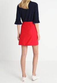 Jupe rouge au design ajusté, coupe droite et fermeture éclair au dos. Associée à un haut bleu marine sombre avec des manches évasées. Des baskets blanches complètent le look.