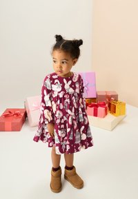 Vinröd klänning med blommönster och långa ärmar, med en gathered midja, matchad med bruna ankellånga stövlar. Färgglada presentaskar i bakgrunden.