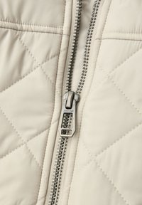 Tissu de veste beige matelassé avec une fermeture éclair argentée sur le devant. La fermeture éclair est équipée d'un curseur et porte le nom de la marque "STREET ONE" gravé.