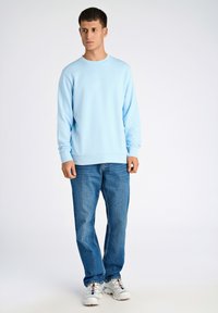 Ljusblå sweatshirt med långa ärmar och rundad nacke, i kombination med blå jeans och vita sneakers. Enkelt mönster med minimala detaljer.