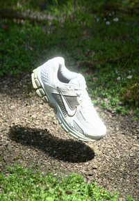 Bela atletska čevlja z mrežasto tkanino, gumijastim podplatom in teksturiranimi detajli. Ima izstopajoč logotip swoosh in obliko zaobljenega nosu.