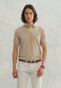 Νεαρός άνδρας με μαλλιά μέχρι τους ώμους, φορώντας μπεζ κοντομάνικο πουκάμισο, λευκά παντελόνια, καφέ ζώνη και ρολόι στον καρπό, μπροστά από απλό ανοιχτό φόντο.