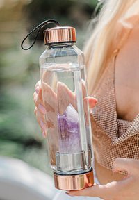 Bouteille d'eau en verre transparent avec un cristal violet à l'intérieur, couvercle en métal doré et base. Dispose d'une sangle noire pour le transport.