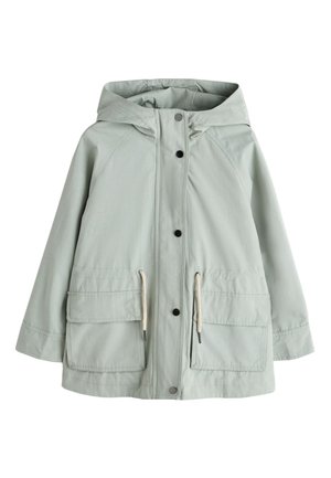 Ung pige iført en lysegrøn hætte-regnjakke, beige strikket cardigan, shorts med blonder og sorte gummistøvler, stående mod en ensfarvet baggrund.