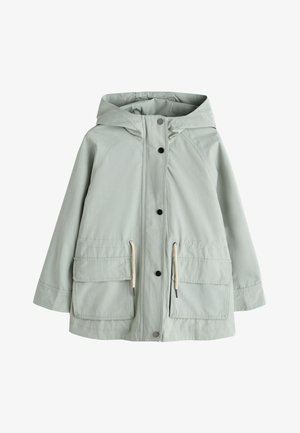 Ung pige iført en lysegrøn hætte-regnjakke, beige strikket cardigan, shorts med blonder og sorte gummistøvler, stående mod en ensfarvet baggrund.