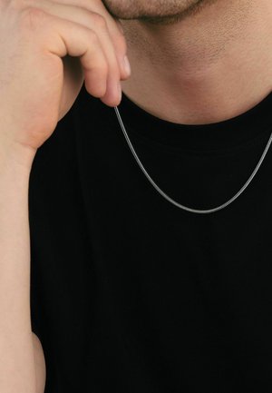 Persona con camisa negra sostiene un collar de cadena delgada de plata cerca de su barbilla, contra su cuello y pecho.