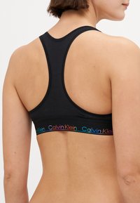 Svart sports-BH med racerback-design, med en regnbue "Calvin Klein" logo-bånd nederst og glattere tekstur.