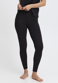 Juodos tamprės iš tamprios medžiagos, su aukštu juosmeniu ir prigludusiu dizainu, pabrėžiančiu kojas. Modelis basas.
