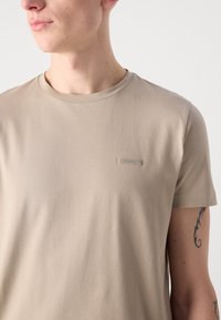 Μπεζ κοντομάνικο μπλουζάκι από μαλακό ύφασμα με στρογγυλή λαιμόκοψη και μικρή επώνυμη ετικέτα στην αριστερή πλευρά του στήθους. Λείος υφή.