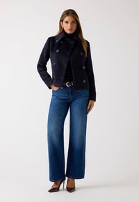 Abrigo de lana combinado en color navy con botones de doble botonadura, acompañado de unos vaqueros de denim azul de pierna ancha y zapatos de tacón oscuros sobre un fondo liso.